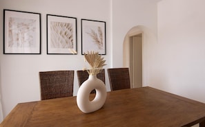 A dining table with a white vase and dried plants, surrounded by wicker chairs. Three framed artworks are hung on the wall above. - Nerina Urban Residence (Paphos)