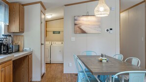 A compact kitchen with a coffee maker, toaster, and a sign indicating a laundry area. A dining table with chairs is positioned near a wall with a beach painting. - Poe's Paradise: Soundside 3 Bedrooms 2 Full Baths - close to beach (Emerald Isle)