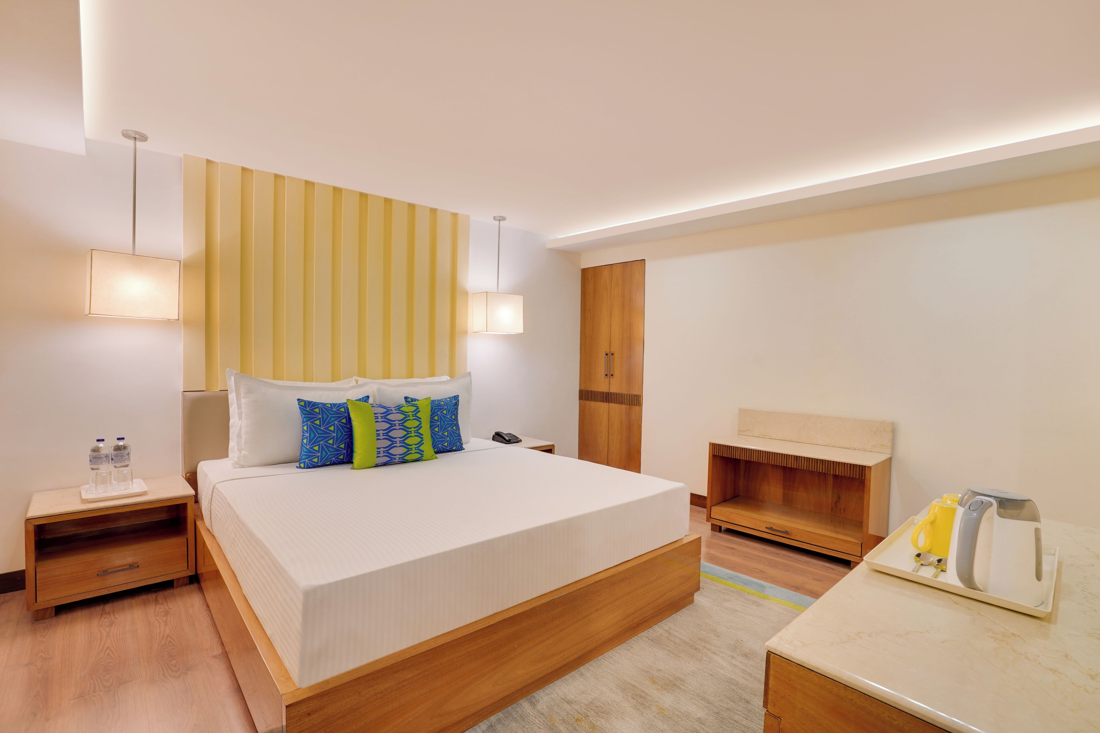 A modern hotel room with a large bed, bedside tables, a wooden wardrobe, and a desk with a kettle and cups.