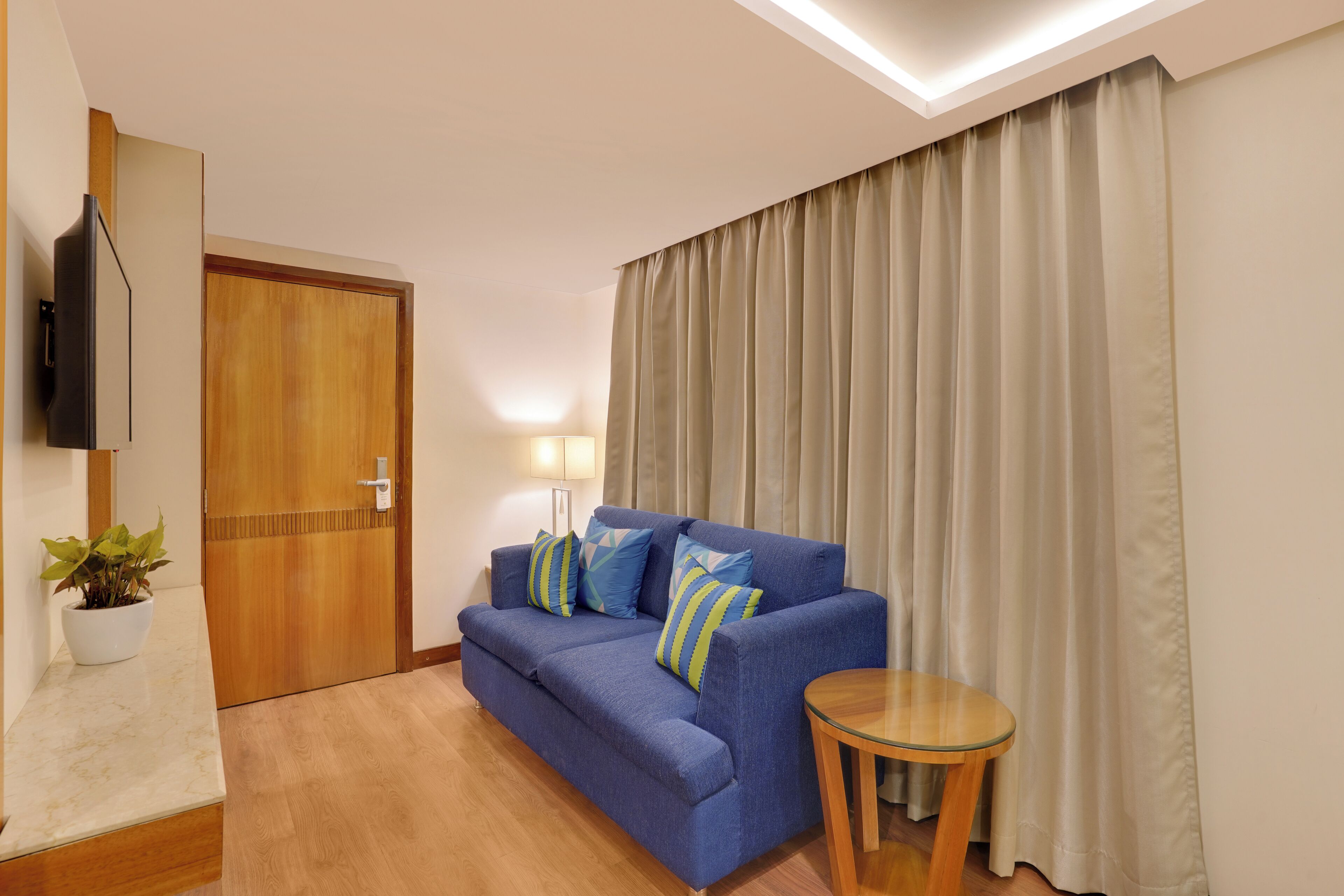 A living room with a sofa, a small round table, a TV mounted on the wall, and a door with a wooden frame.