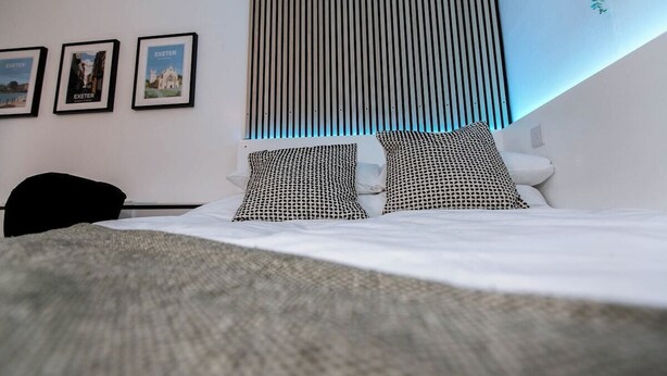 A hotel room with a bed, black and white patterned pillows, a black chair, and three framed pictures on the wall. - Studio With Parking, By Uni/Hosp (Exeter)