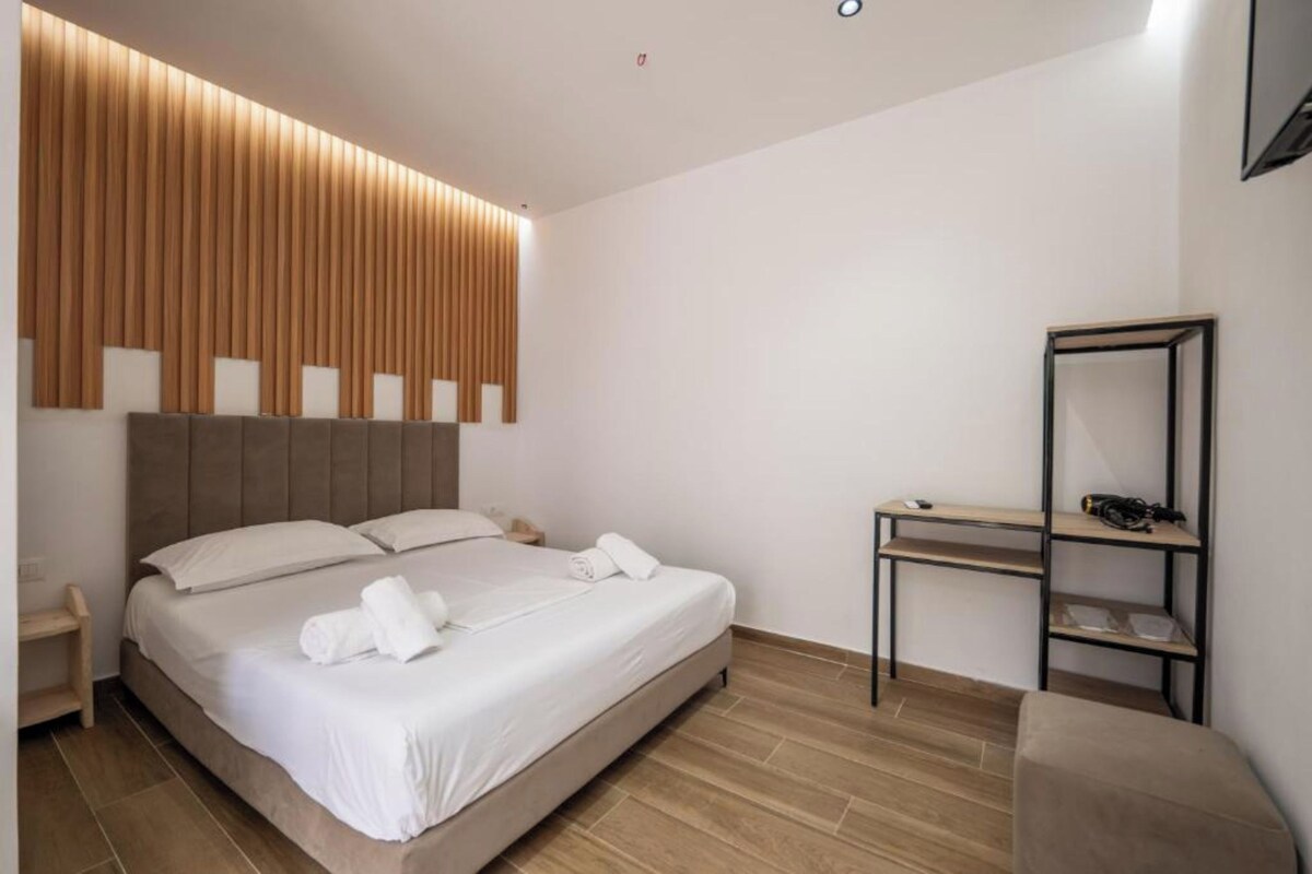 A modern hotel room with a large bed, a wooden panel wall, a small desk, and a chair.
