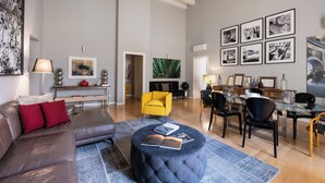 Un vaste espace de vie comprenant un canapé moderne, un pouf rond sur lequel sont posés des livres, un fauteuil jaune vif et une table à manger avec des chaises.