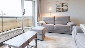 Ein modernes Wohnzimmer mit einer Couch, einem Hocker und einem Holztisch. Ein großes Fenster bietet Blick auf einen Strand.