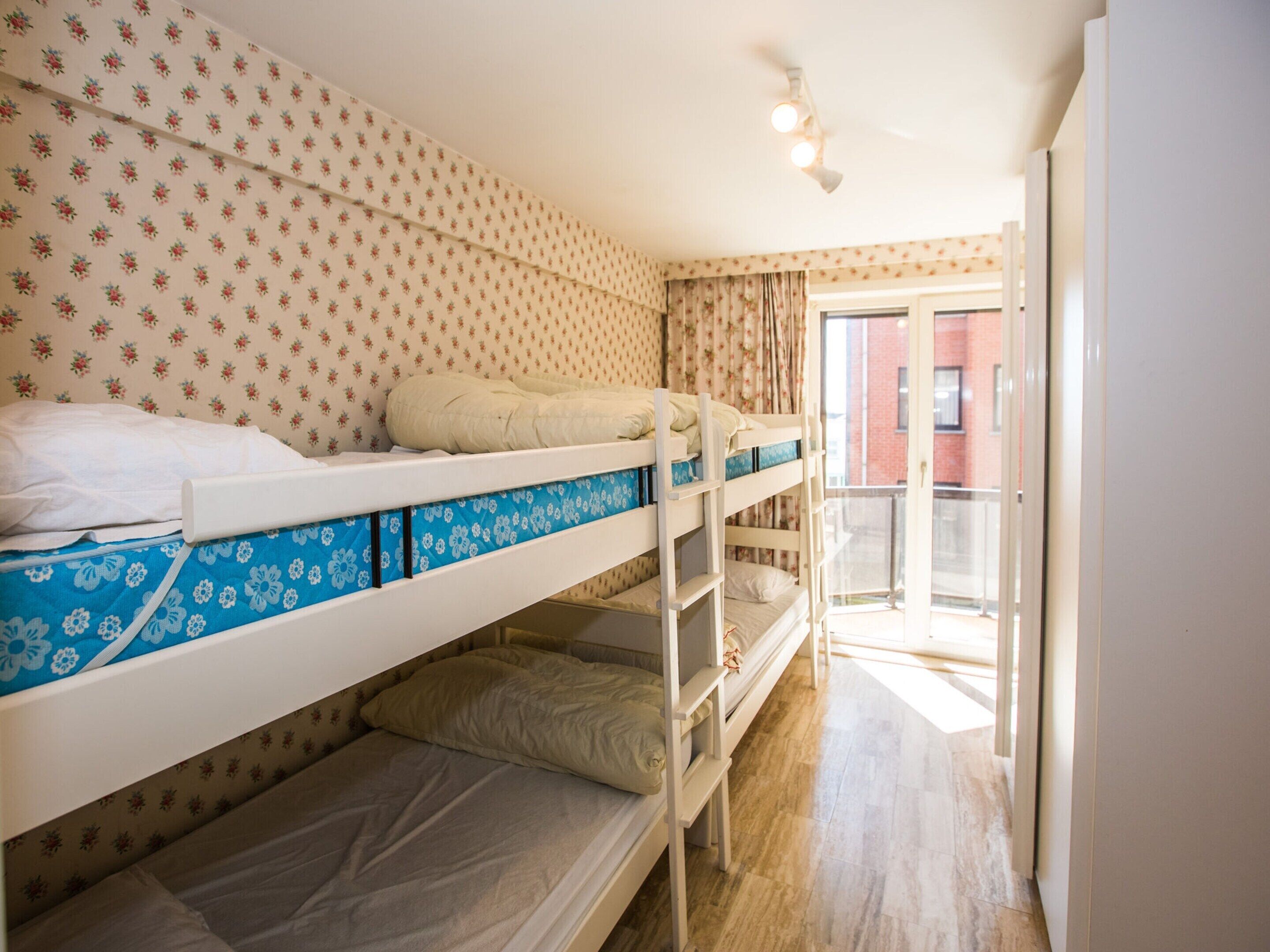 Een studentenkamer met stapelbedden, bloemenbehang en uitzicht op een gebouw door een glazen deur.