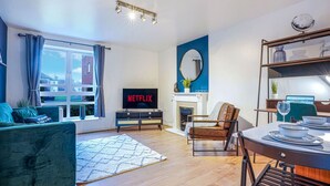 Ein modernes Wohnzimmer mit einem Fernseher, der 'NETFLIX' zeigt, einem Kamin, einem Essbereich mit gedecktem Tisch und einem großen Fenster mit Vorhängen.