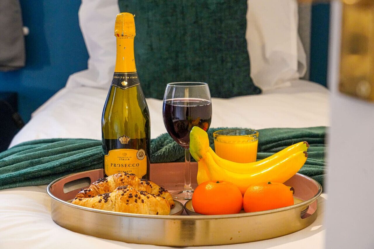 A breakfast tray with croissants, bananas, oranges, a bottle of Prosecco, and a glass of red wine on a bed with a green blanket.
