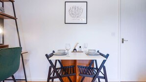 Ein Essbereich mit weißem Tisch, gedeckt für vier Personen, mit je zwei Stühlen an jeder Seite. An der Wand hängt ein botanischer Kunstdruck, und ein Holzlader lehnt an der Wand.