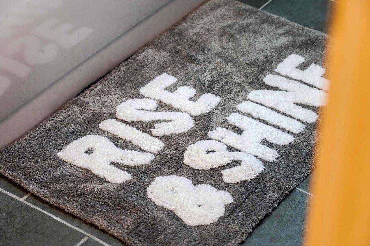 A doormat with the words "RISE & SHINE" in white letters on a grey textured surface.