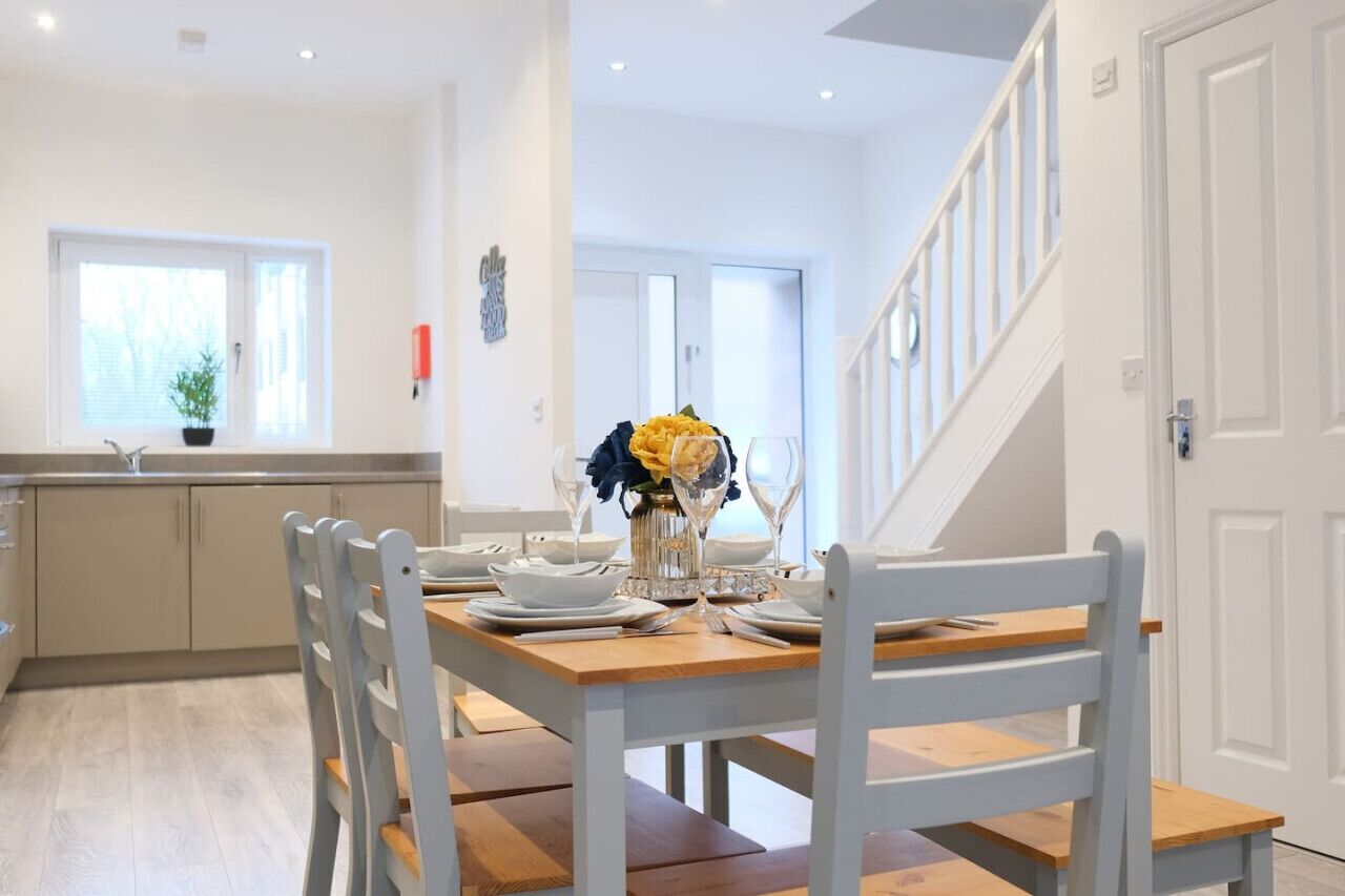 A dining area with a wooden table set for four, white chairs, and a vase with yellow flowers. A kitchen with a window and a plant is visible in the background.