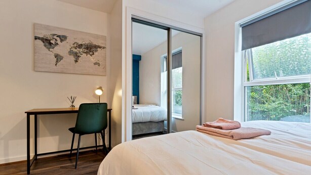 A bedroom with a bed, a desk, a chair, a world map artwork, and a view of greenery outside. - Stylish Apartment in City Centre with Free Parking (Chelmsford)