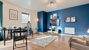 A modern living room with a blue accent wall, a dining area with a table and chairs, a sofa, and a TV mounted on the wall. - Stylish Apartment in City Centre with Free Parking (Chelmsford)