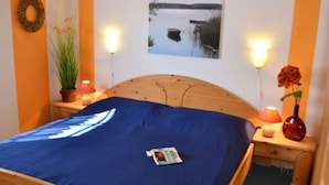 Un dormitorio con una cama de madera, un cuadro enmarco de un lago, una planta en maceta y un florero con flores.