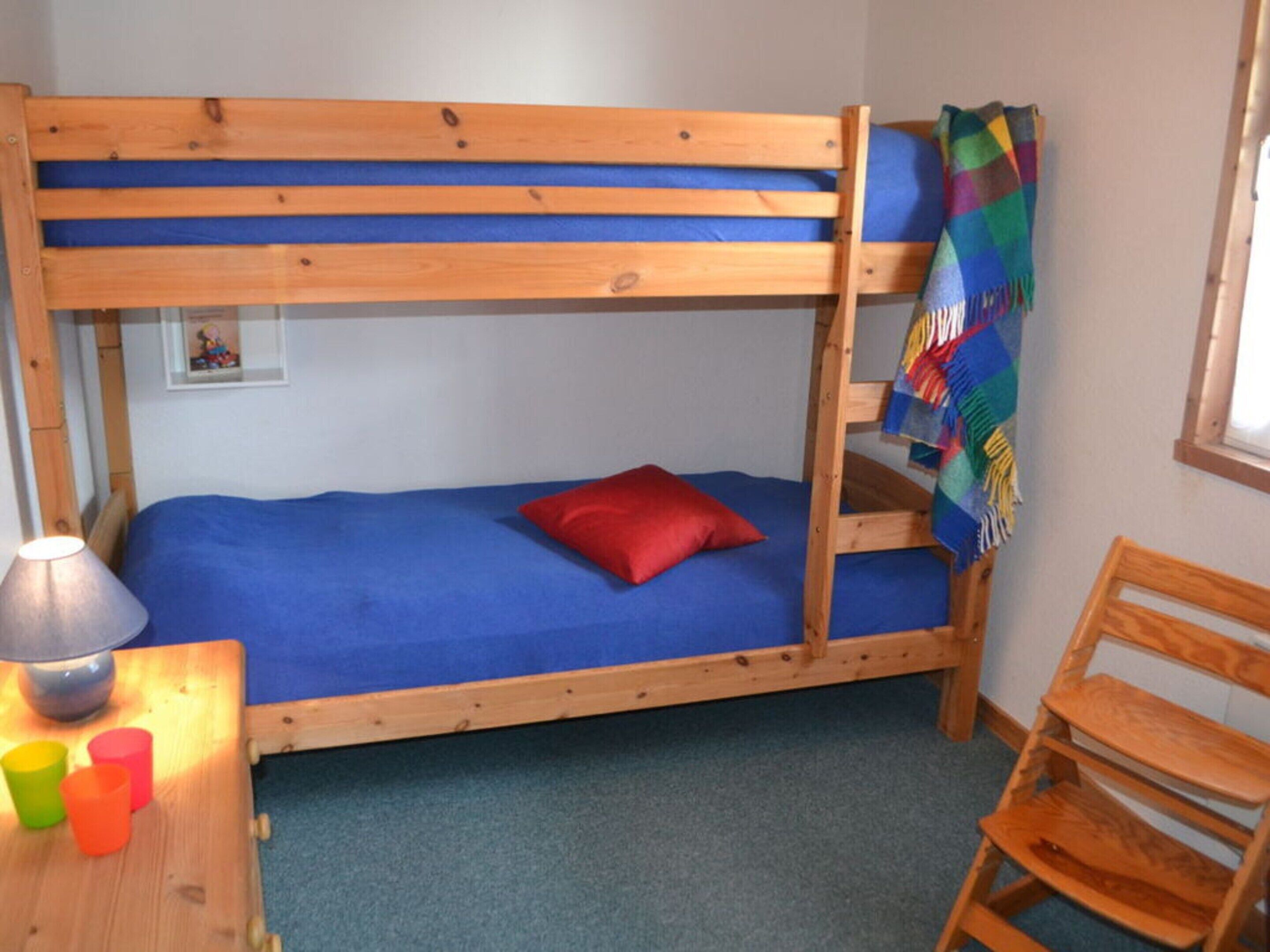 A bunk bed with a blue mattress and a red pillow, a wooden chair, a small table with a lamp and colorful cups, and a colorful blanket on the right side.