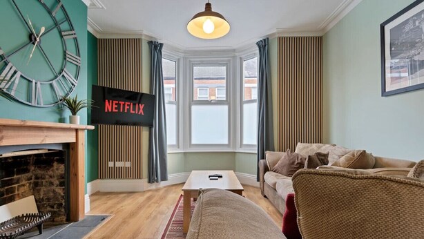 A living room with a fireplace, a large clock, a television displaying 'NETFLIX', a sofa, and a window with curtains.