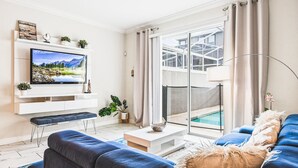 Ein modernes Wohnzimmer mit einem Flachbildfernseher, einer Couch, einem Couchtisch und Blick auf einen Swimmingpool durch eine Glastür.