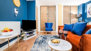A modern living room with a blue wall, a flat-screen TV, a blue chair, an orange sofa, and a coffee table with snacks and drinks. - Navy Retreat - Free Parking, Fast WiFi & SmartTV (Derby)