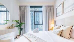 A modern hotel room with a large bed, a round mirror, a potted plant, and a view of tall buildings through the window. - Charming 1BR Apartment in Creek Edge Tower 1, Dubai Creek Harbor by Deluxe Holiday Homes (Dubai)