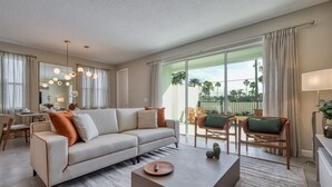 A modern living room with a sofa, coffee table, and chairs. There is a large window with a view of palm trees and a balcony. - Last Minute Disney Getaway! Private Pool & Themed Bedrooms. Book Now! #104041 (Orlando)