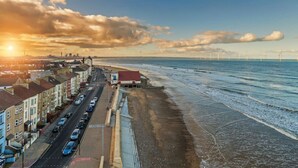Una carretera costera con edificios a un lado y una playa al otro, y aerogeneradores en la distancia.