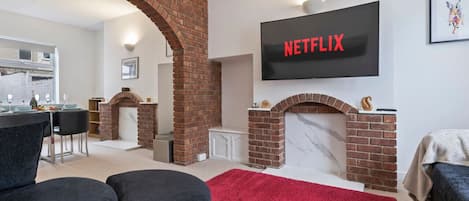 A living room with a brick fireplace, a flat-screen TV mounted above it, and a red rug on the floor.