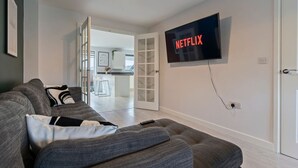 A modern living room with a grey sofa, a mounted TV displaying the Netflix logo, and a door leading to a kitchen area.