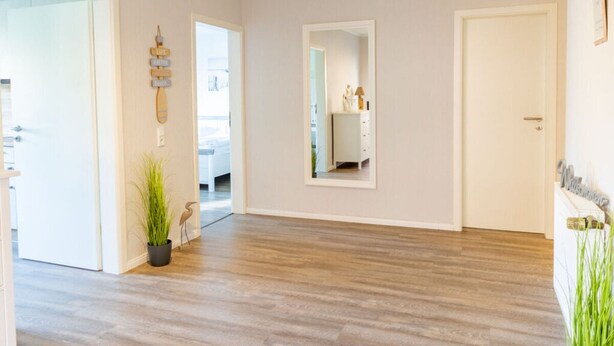 A spacious hallway with wooden flooring, a white door, and a mirror reflecting a bathroom. - Holiday Home Tidenkieker (Nordstrand)