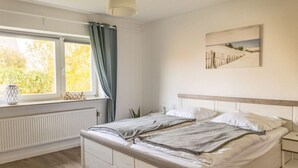 A bedroom with a bed, a bedside table, a window with curtains, a radiator, and a painting on the wall. - Holiday Home Tidenkieker (Nordstrand)
