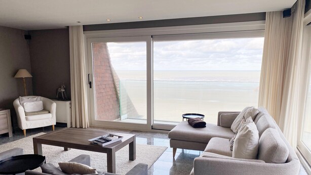 A modern living room with a sofa, armchair, and coffee table, a lamp, and a view of the sea through large windows. - Apartment in Nieuwpoort by Sandy Beaches (Nieuwpoort)