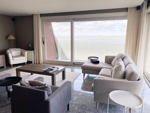 Ein modernes Wohnzimmer mit einer Couch, einem Sessel und einem Couchtisch, einer Lampe und Meerblick durch große Fenster.