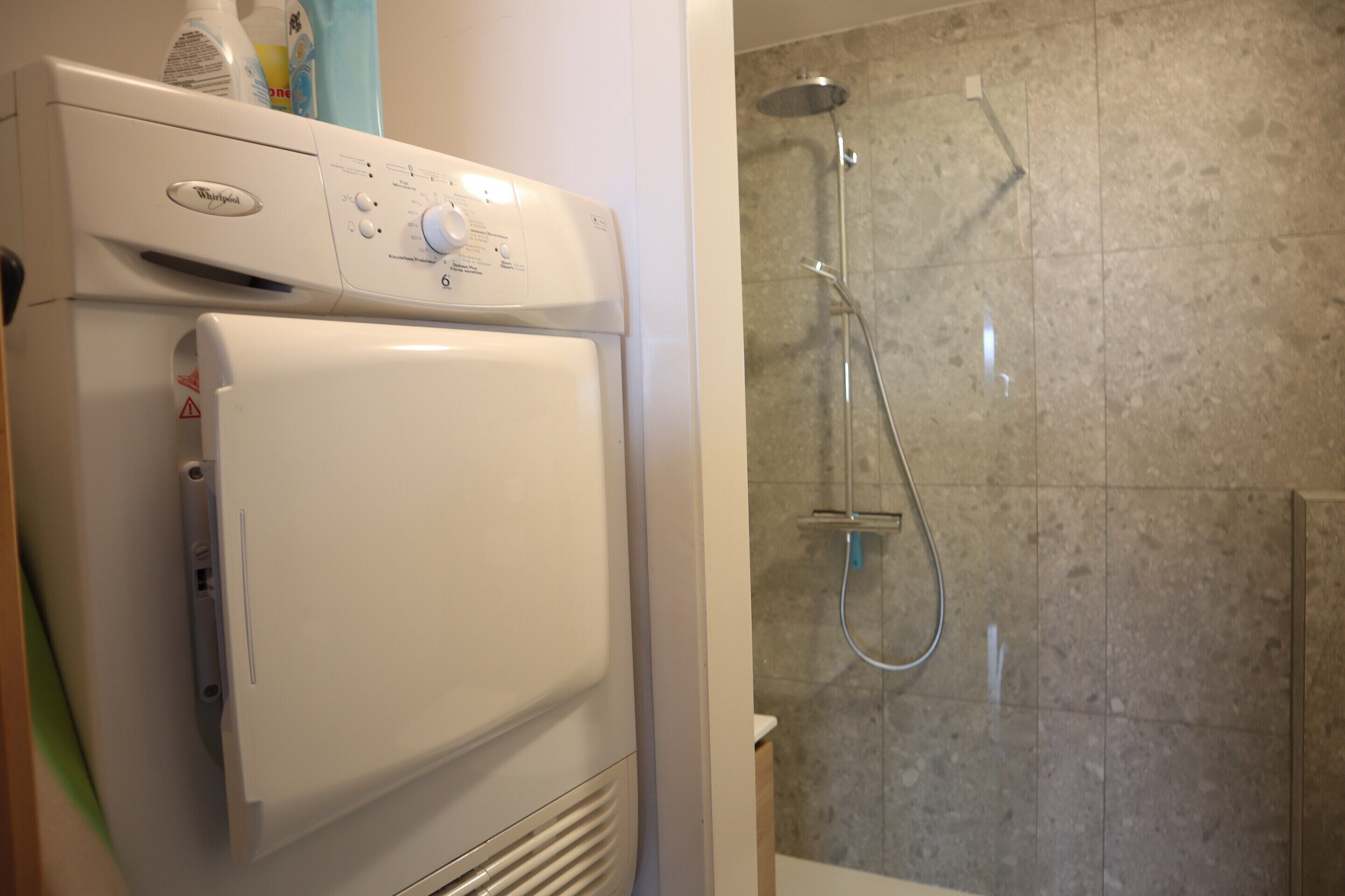 A front-loading washer and dryer unit with a control panel, a shower with a handheld showerhead, and a tiled shower wall.