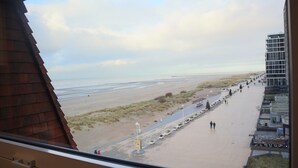 A view from a window overlooking a beach with a paved walkway, a few people, and a building with a sign that reads "RECYCLE CLING". - Apartment in Nieuwpoort by Sandy Beaches (Nieuwpoort)