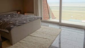 A bedroom with a bed, a nightstand, a window with a beach view, and a carpet. - Apartment in Nieuwpoort by Sandy Beaches (Nieuwpoort)