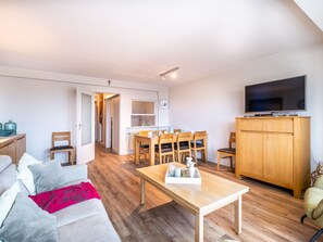 A modern living room with a sofa, a coffee table, a dining table with chairs, a TV on a wooden cabinet, and a hallway leading to a kitchen. - Apartment Near Belgian Coast With Sea View (Koksijde)