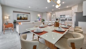 A modern kitchen with white cabinets, a central island, and a dining area with a long table set for six. - Last Minute Disney Getaway! Private Pool & Themed Bedrooms. Book Now! #104175 (Orlando)