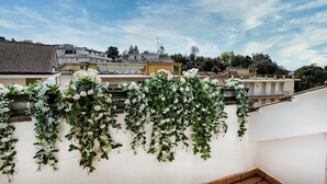 A rooftop garden with white and pink flowers, a clear blue sky, and residential buildings in the background.
