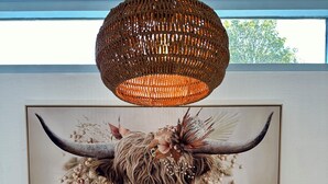 A dining area with a wooden table, a woven pendant light, a framed artwork of a yak, and a vase with dried plants.