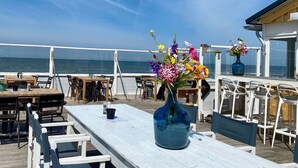 A deck with tables and chairs, a blue vase with flowers, and a view of the ocean.