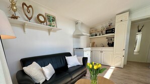 A modern living room with a black leather sofa, white cushions, a wooden coffee table, and a vase of yellow flowers. A kitchen with white cabinets and a microwave is visible in the background.