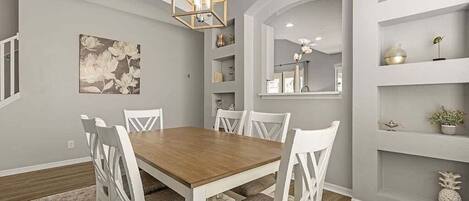 Un comedor con una mesa de madera y sillas blancas, una araña y una pared decorativa con estanterías.