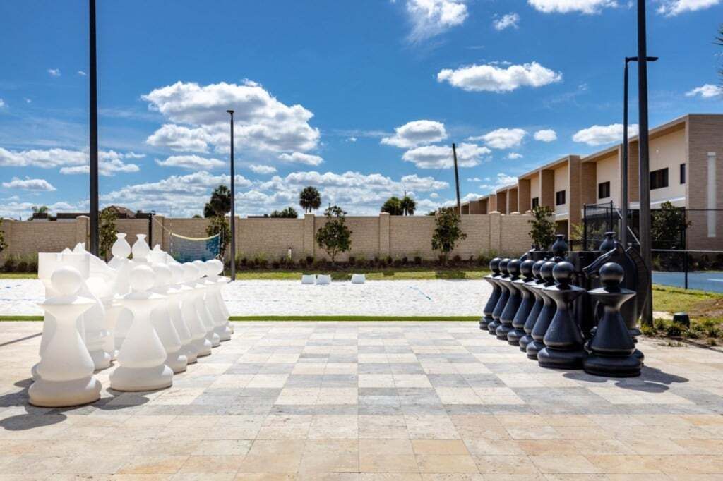 A large outdoor chessboard with white and black pieces, set against a backdrop of a clear sky and a building with a brick wall and windows.