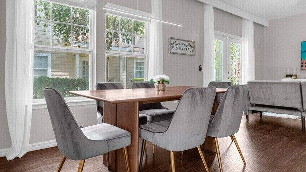 A dining area with a wooden table and grey upholstered chairs, large windows with a view of greenery, and a wall-mounted sign.