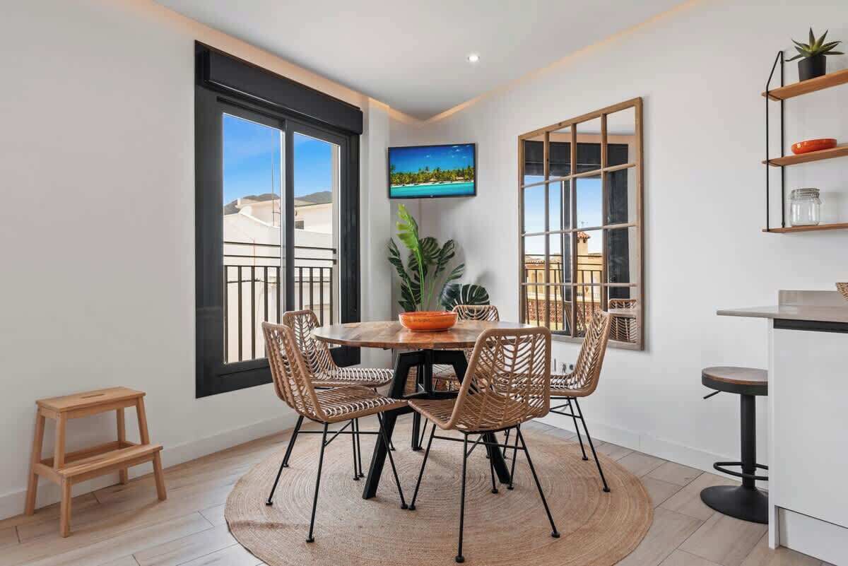 A dining area with a round table, wicker chairs, a small wooden stool, and a large window with a view of buildings.