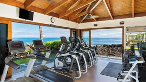 A gym with treadmills and exercise equipment, large windows with a view of the ocean, and a wooden ceiling.