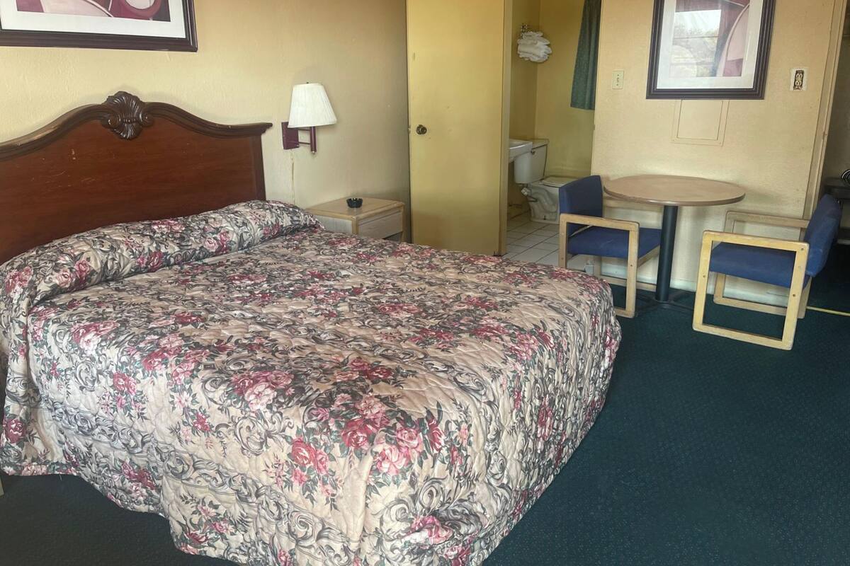A hotel room with a bed, a small table with two chairs, a lamp, and a bathroom visible through an open door.
