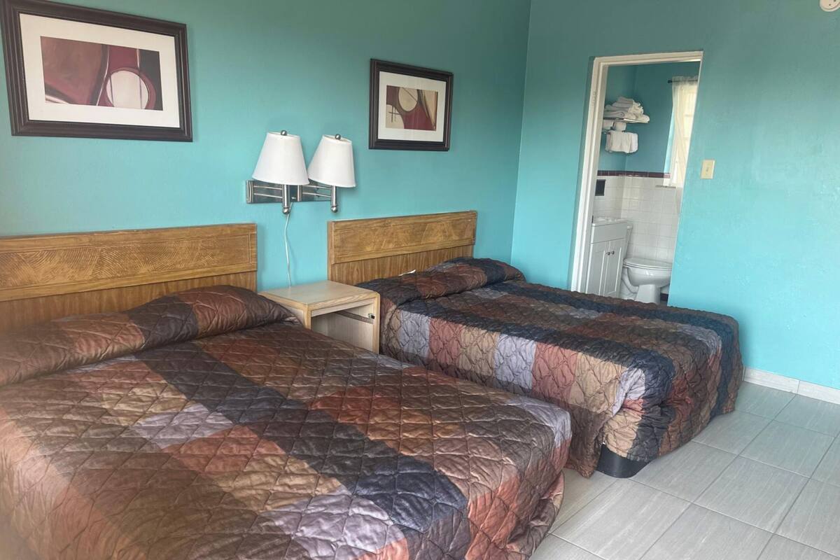 A hotel room with two beds, a nightstand, two wall-mounted lamps, and a bathroom visible through an open door.