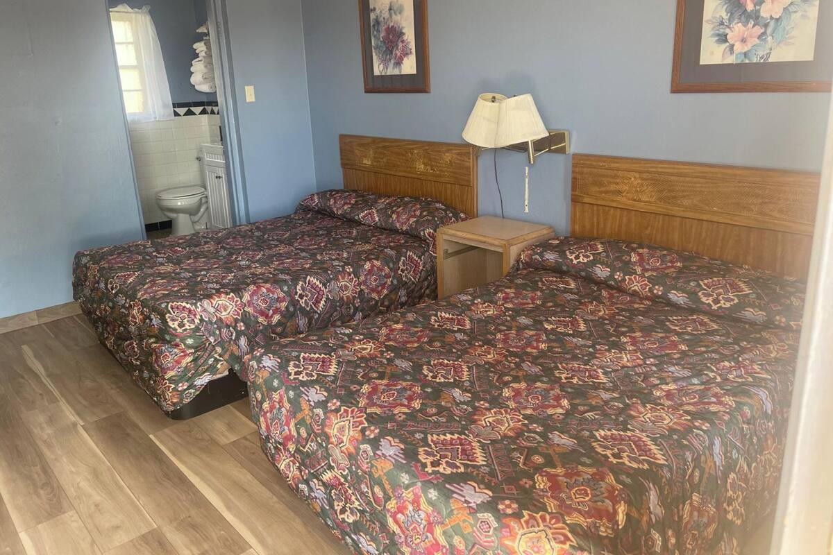 A room with two beds, a wooden headboard, a bedside table with a lamp, a mirror, and a toilet.