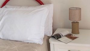Una cama bien hecha con ropa blanca, cabecera roja, una lámpara con base texturizada y una revista sobre una mesita de noche.