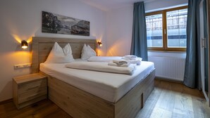 A neatly made bed with pillows and folded towels, a bedside table with drawers, a wall-mounted light, and a window with curtains.