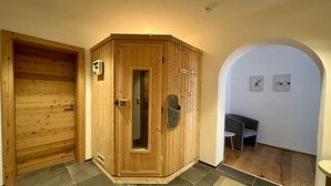 Uma sauna de madeira com porta, uma mesa pequena e um banco em um espaço com piso ladrilhado e parede branca com obras de arte.